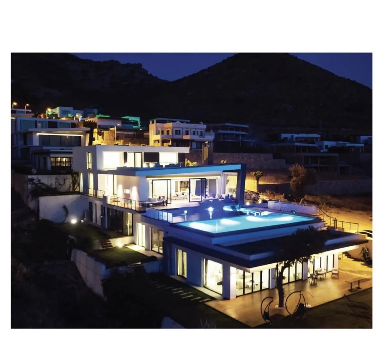 Marinhill Luxury Villas seen as a whole at dusk — a multi-level modern villa on the hillside with lit interiors, wide terraces and an illuminated infinity pool