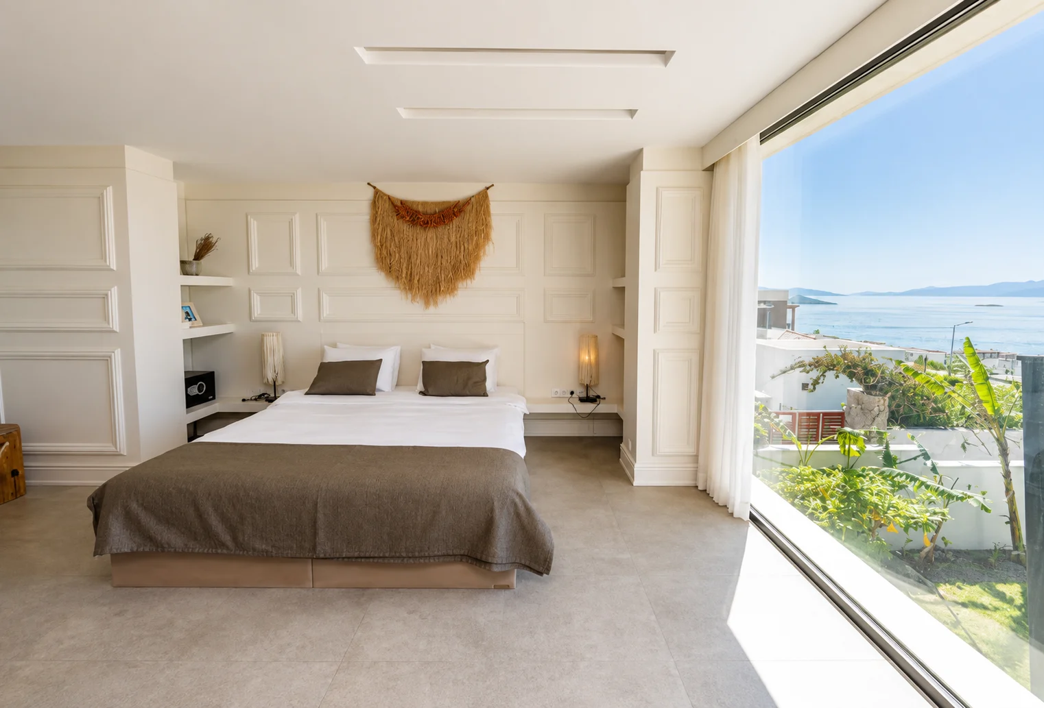 Sea View room with a wide double bed in cream tones and floor-to-ceiling glass opening onto the Aegean and the islands